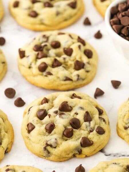Homemade chocolate chip cookies lined up on a kitchen countertop with some loose chocolate chips
