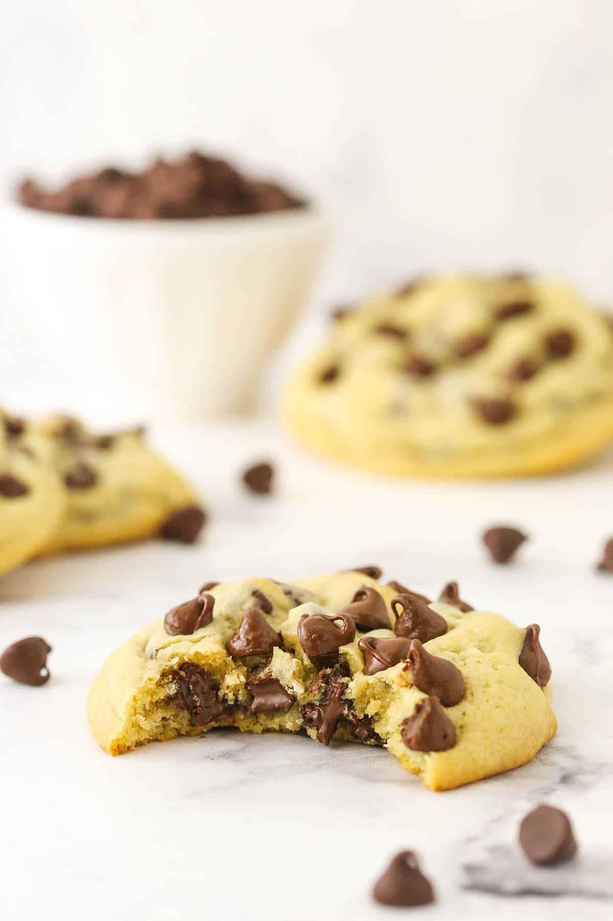 A soft and chewy chocolate chip cookie on a marble kitchen countertop with a big bite taken out of it to reveal a bunch of melty chocolate chips in the center