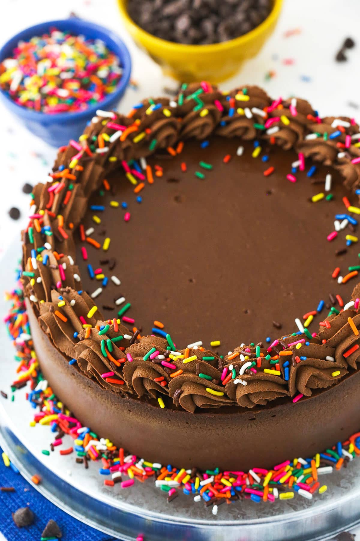 A chocolate cheesecake on a cake platter with rainbow sprinkles on top