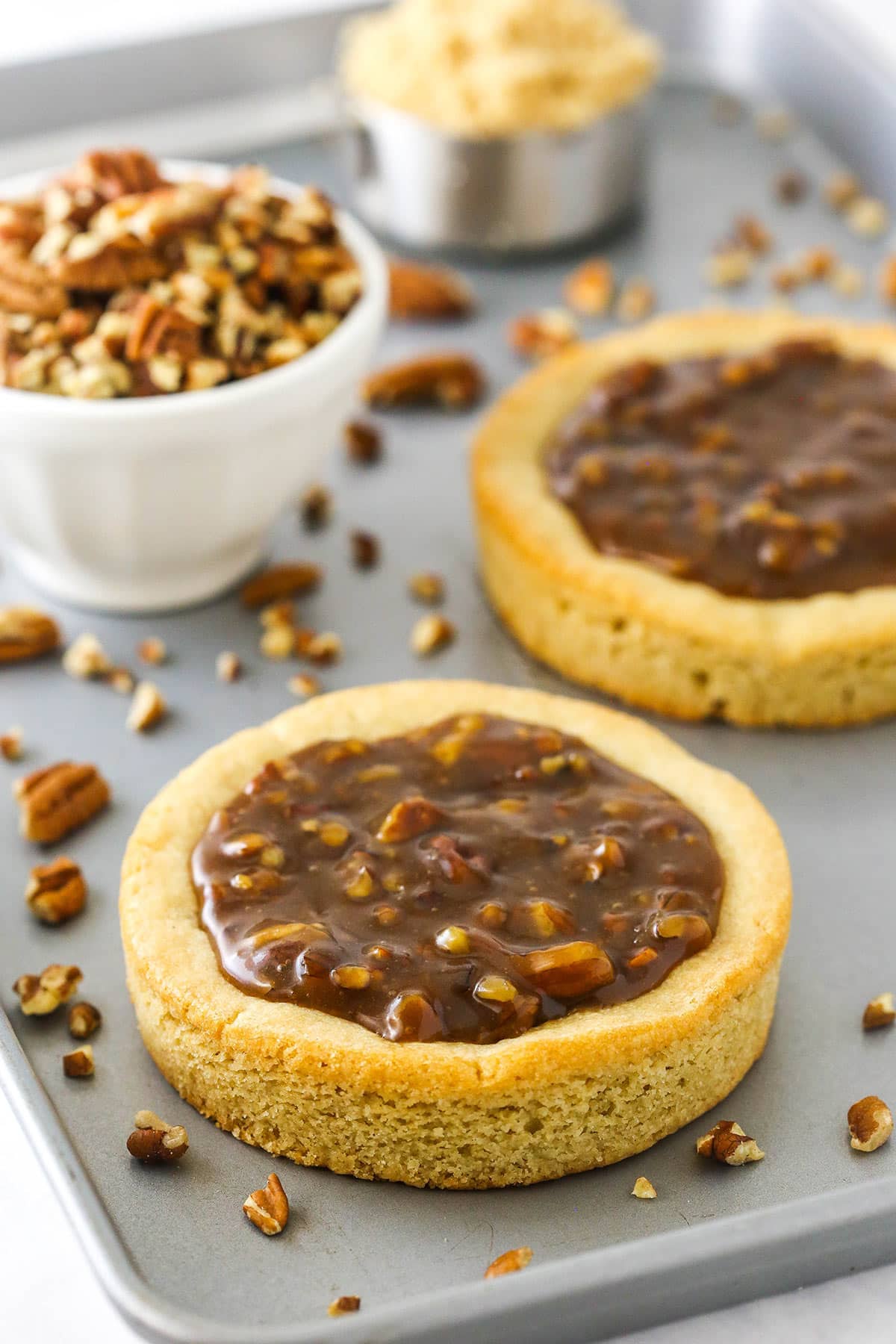 Two pecan pie cookies on a baking sheet beside a bowl of chopped pecans