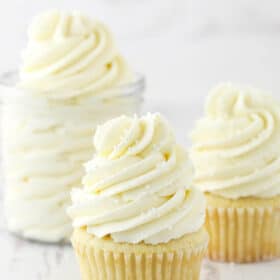 two frosted cupcakes and jar of frosting on white background