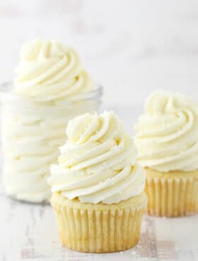 two frosted cupcakes and jar of frosting on white background