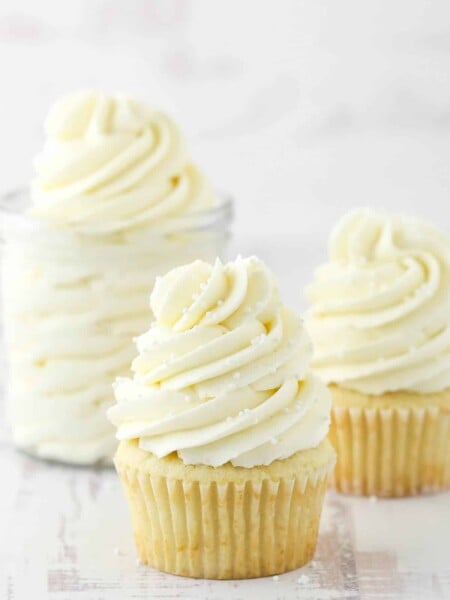 two frosted cupcakes and jar of frosting on white background