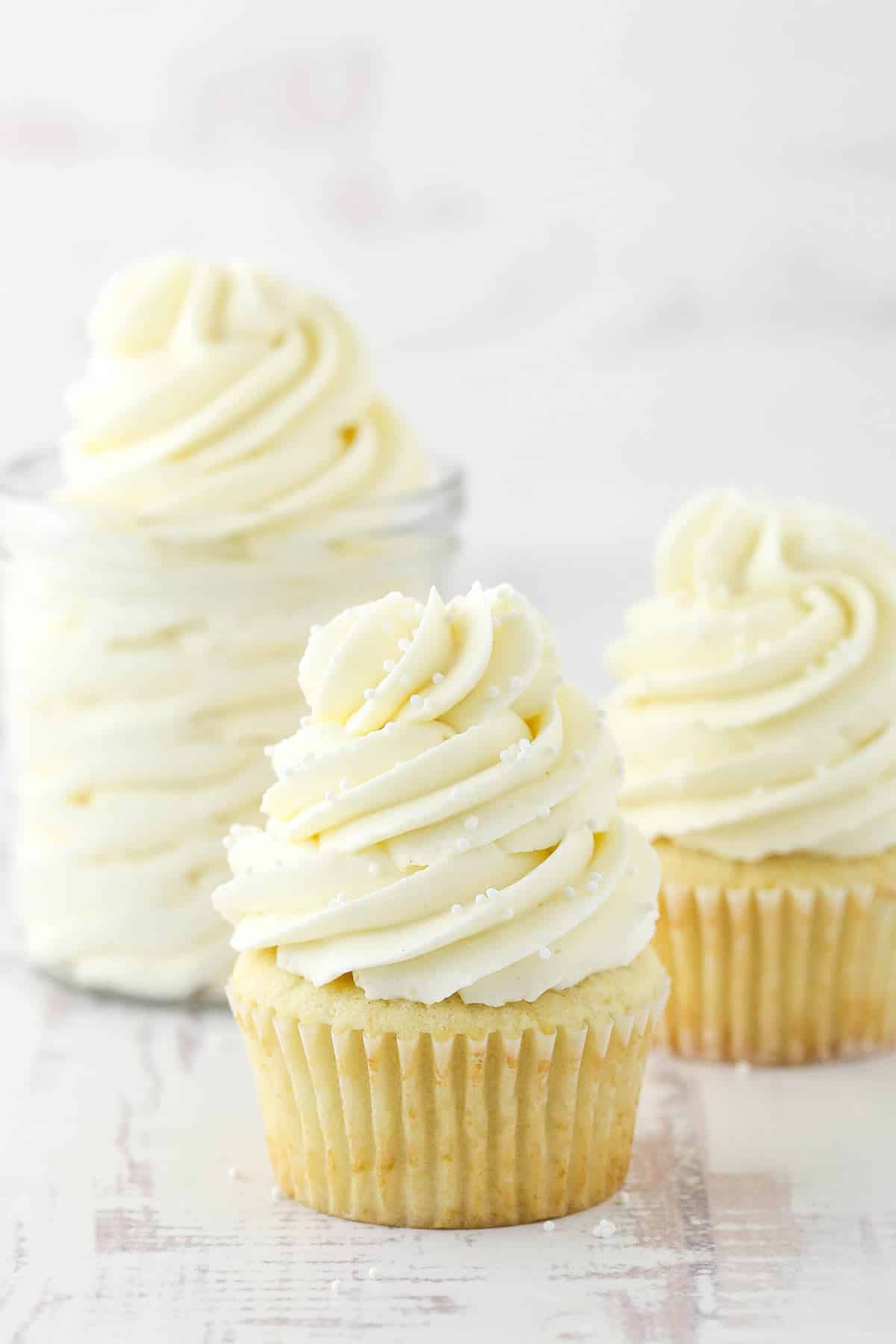 two frosted cupcakes and jar of frosting on white background