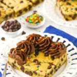 A big corner slice of cookie cake on a plate with chocolate chips and sprinkles in small dishes behind it