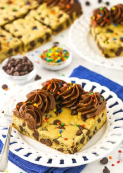 A big corner slice of cookie cake on a plate with chocolate chips and sprinkles in small dishes behind it