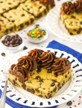 A big corner slice of cookie cake on a plate with chocolate chips and sprinkles in small dishes behind it