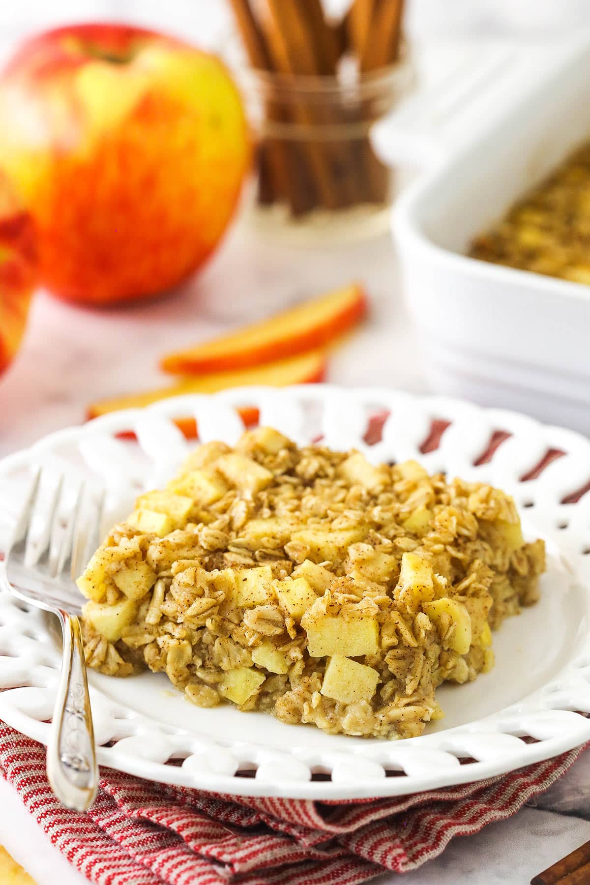 Filled with pops of fresh apple and warm, cozy spices, Baked Apple Cinnamon Oatmeal is a fall must-have that hits the spot all year long! One portion of baked oatmeal on a plate with the remaining oatmeal bake in the background