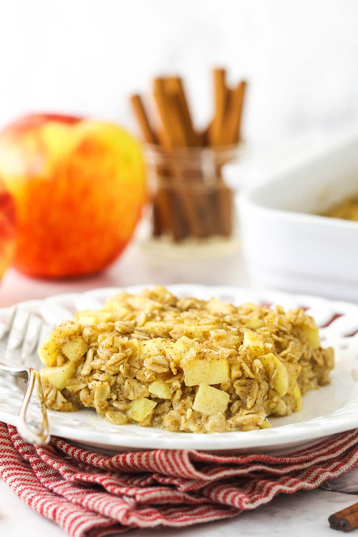 A plate holding a piece of baked oatmeal with an apple and a jar of cinnamon sticks behind it