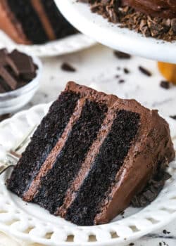 slice of Devil's Food Cake on white plate with another slice and bowl of chocolate in the background