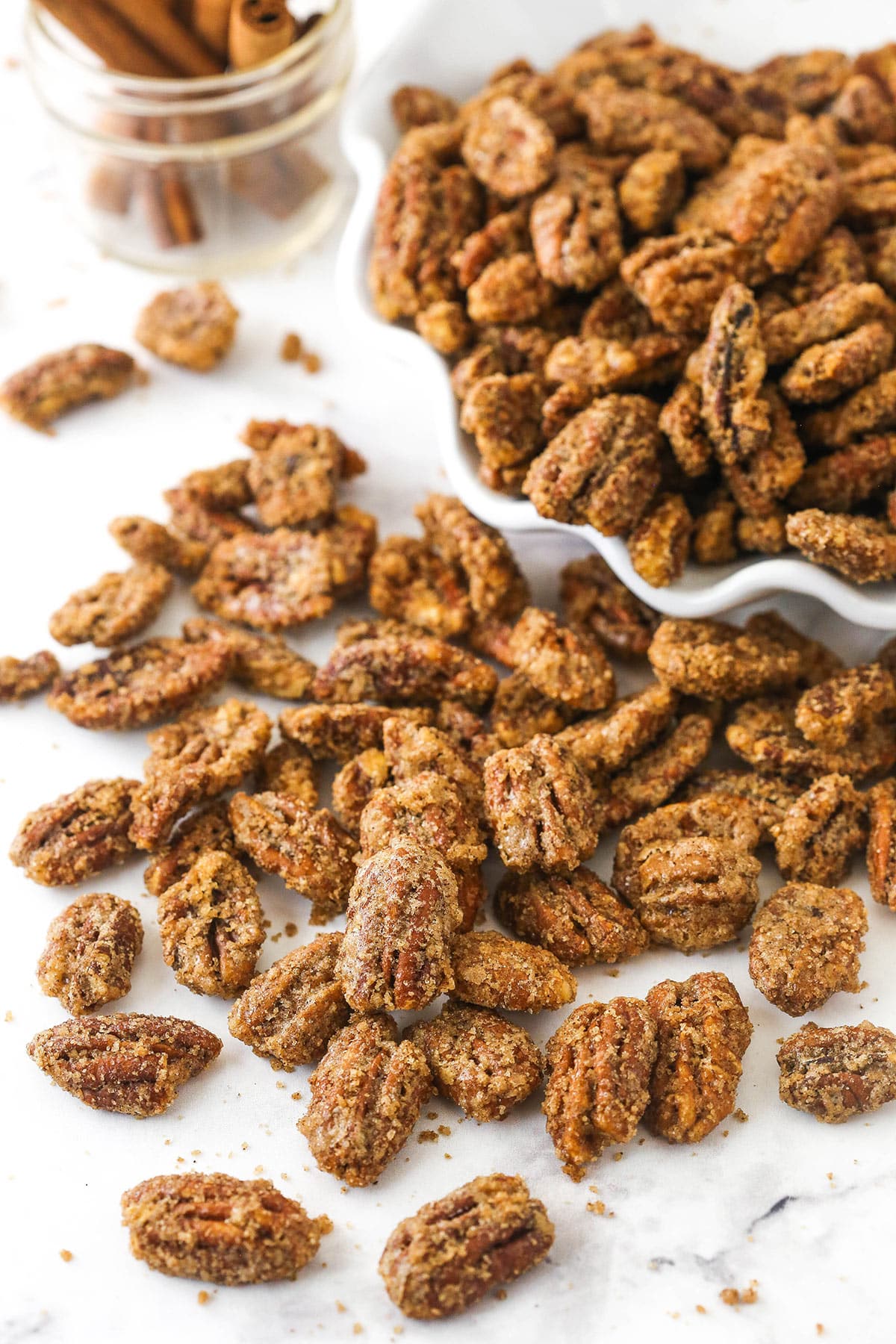 Covered in a cinnamon sugar coating and caramelized in the oven, these crunchy candied pecans will quickly become your new favorite snack. They're super easy to make, and they stay fresh for weeks! A bunch of candied pecans in a bowl with more spilling out onto a kitchen countertop.