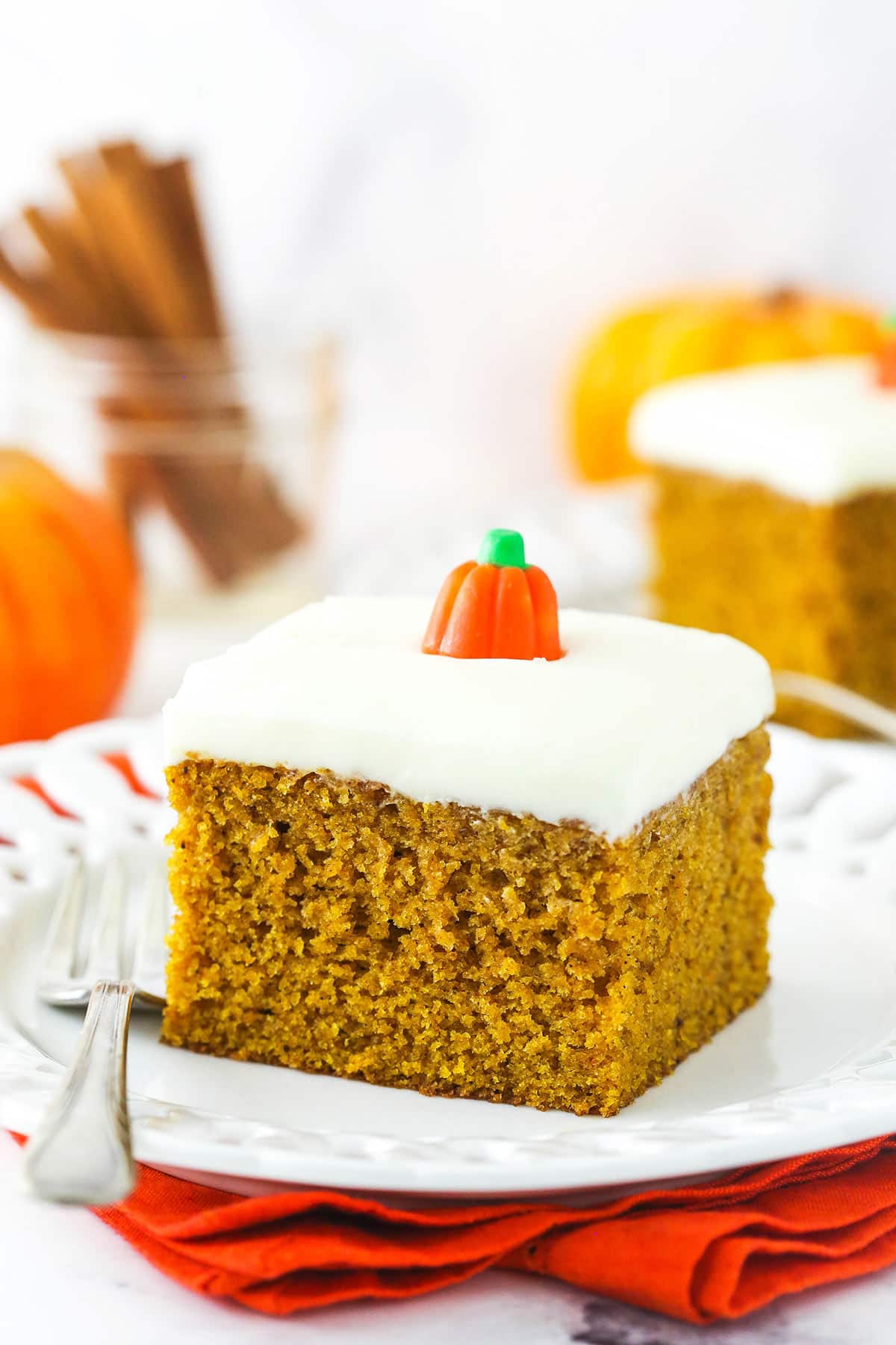 A square slice of pumpkin cake on a plate with cinnamon sticks in a jar behind it.