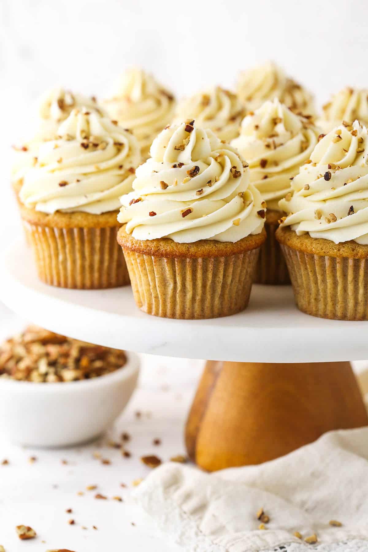 A cake stand holding a batch of pecan pie cupcakes with a bowl of toasted and chopped pecans beside it