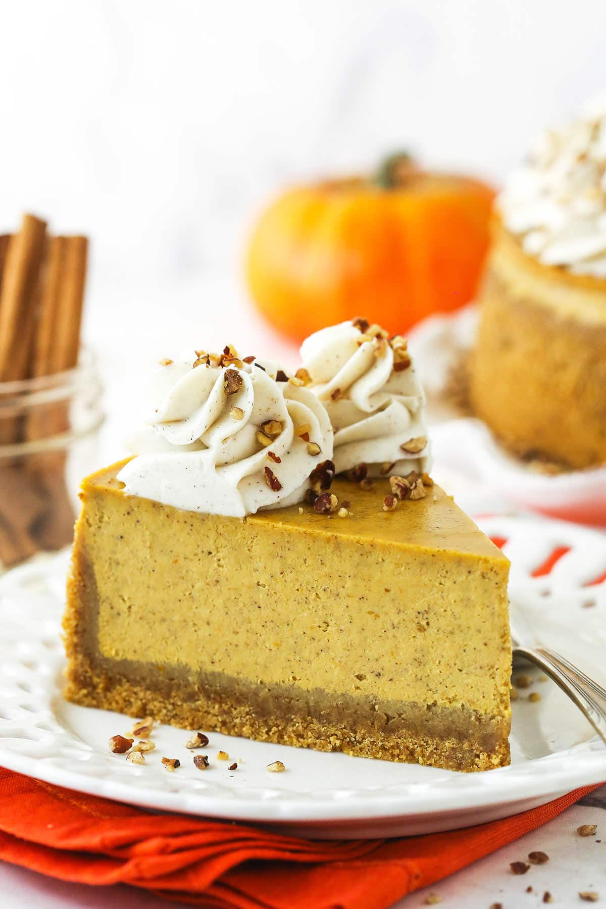 A slice of pumpkin cheesecake on a plate with a pumpkin and a jar of cinnamon sticks behind it