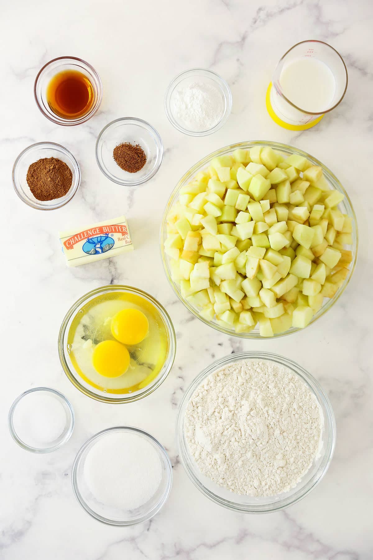 Chopped apples, flour, eggs, butter and the rest of the ingredients arranged neatly on top of a marble kitchen counter