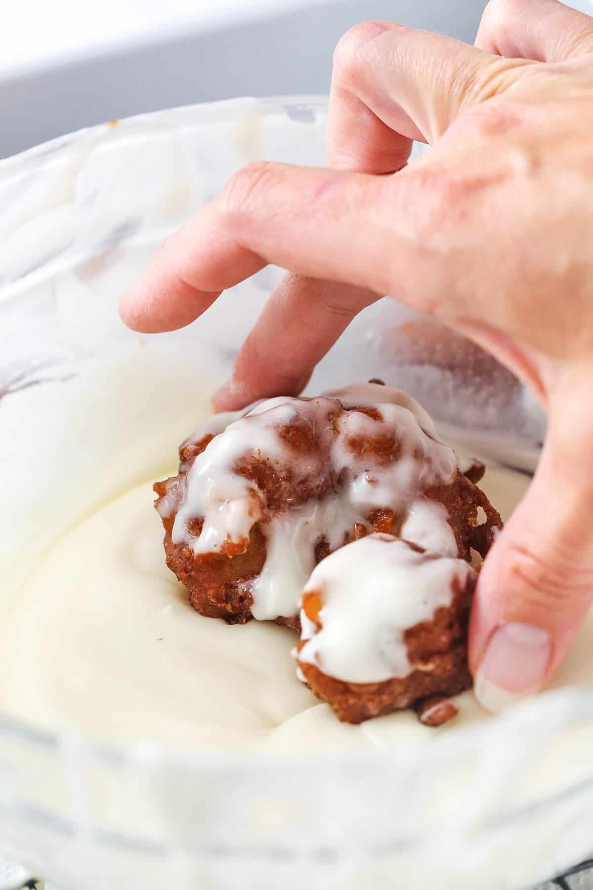 An apple fritter being dipped into a bowl of vanilla glaze