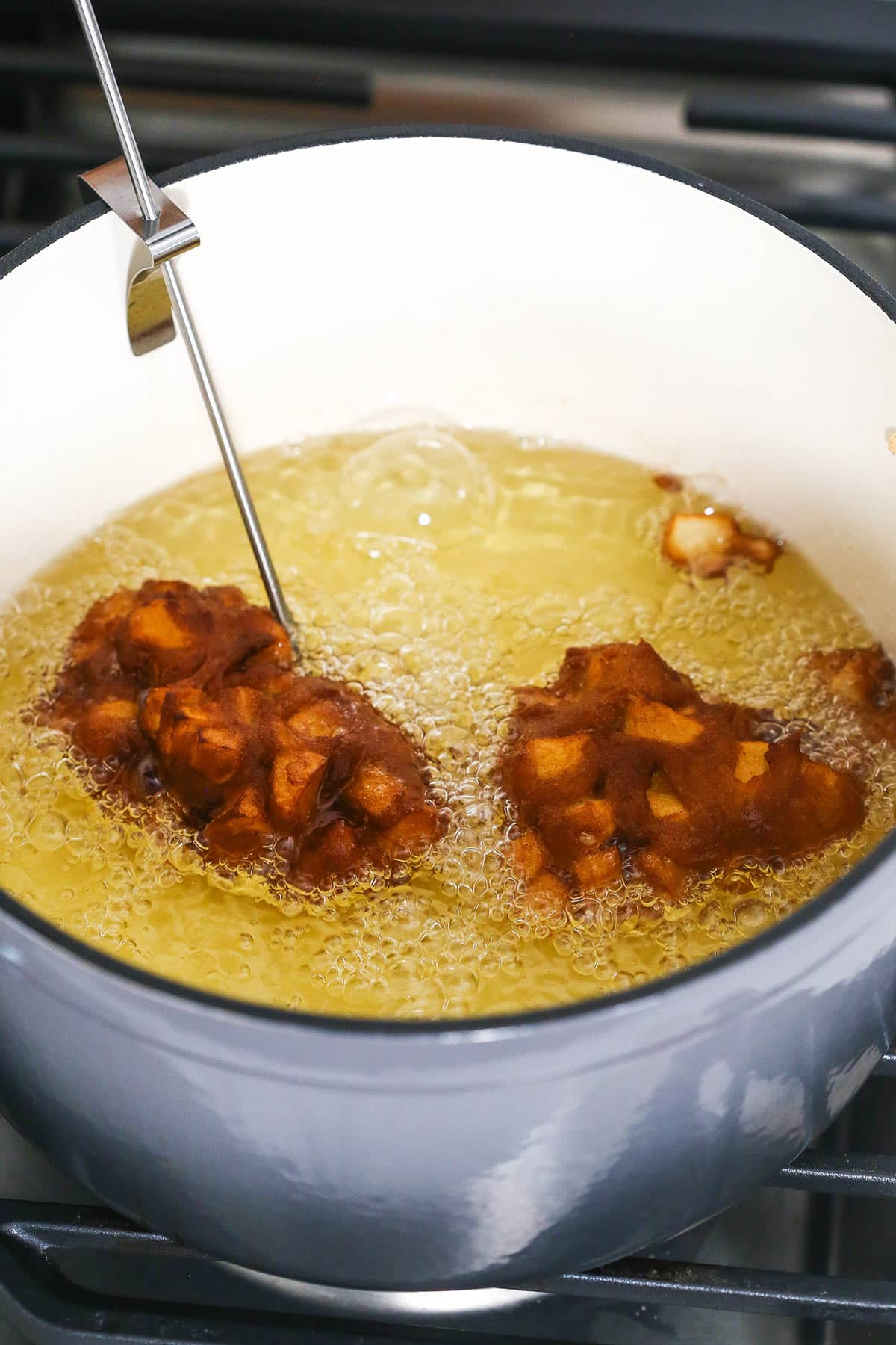 Two apple fritters frying in a pot of oil with a candy thermometer stuck inside