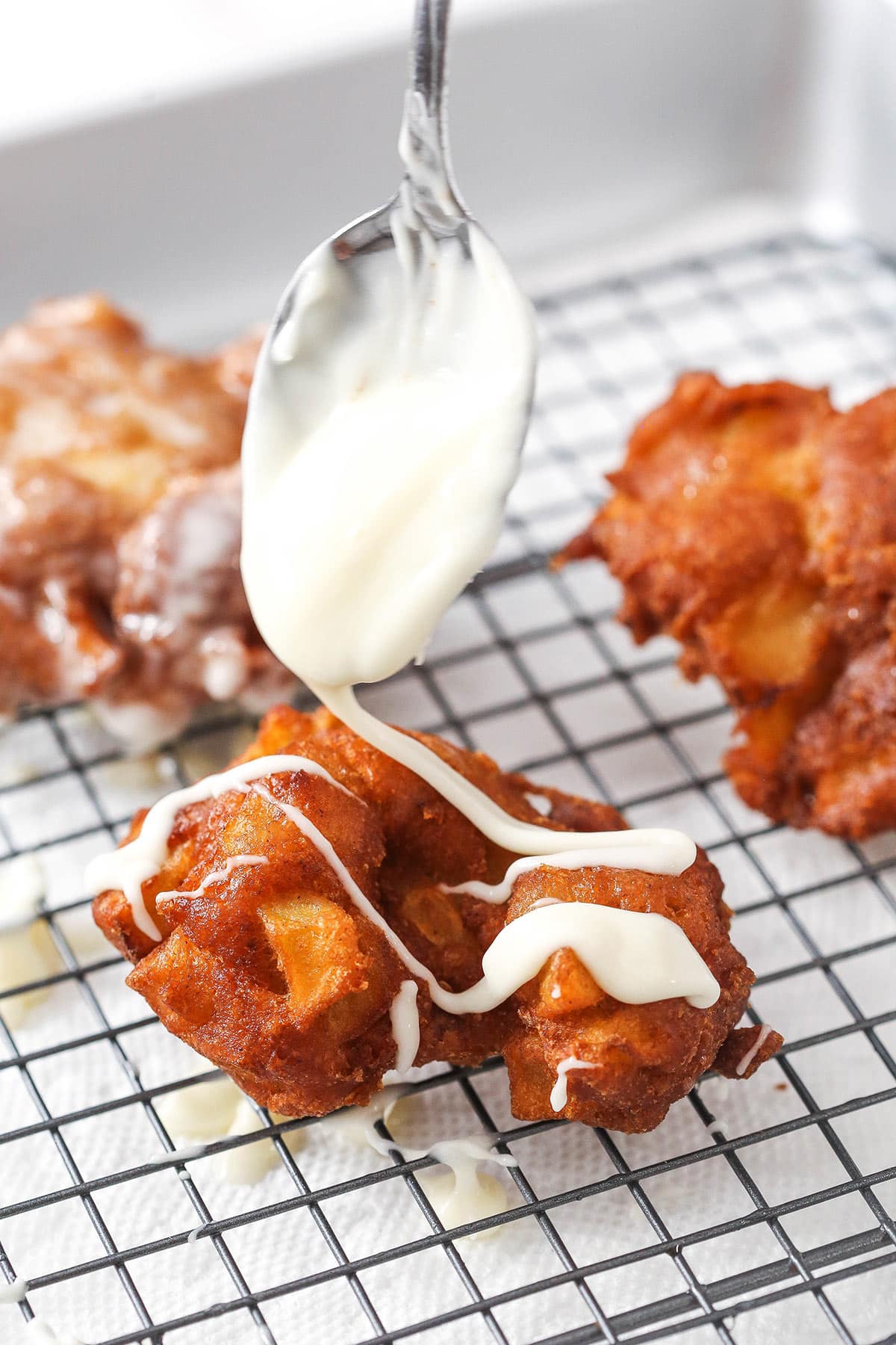 A spoon drizzling some homemade vanilla glaze over an apple fritter on a cooling rack