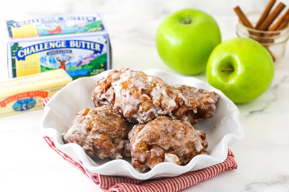 A serving platter full of apple fritters with fresh apples, cinnamon sticks and butter in the background
