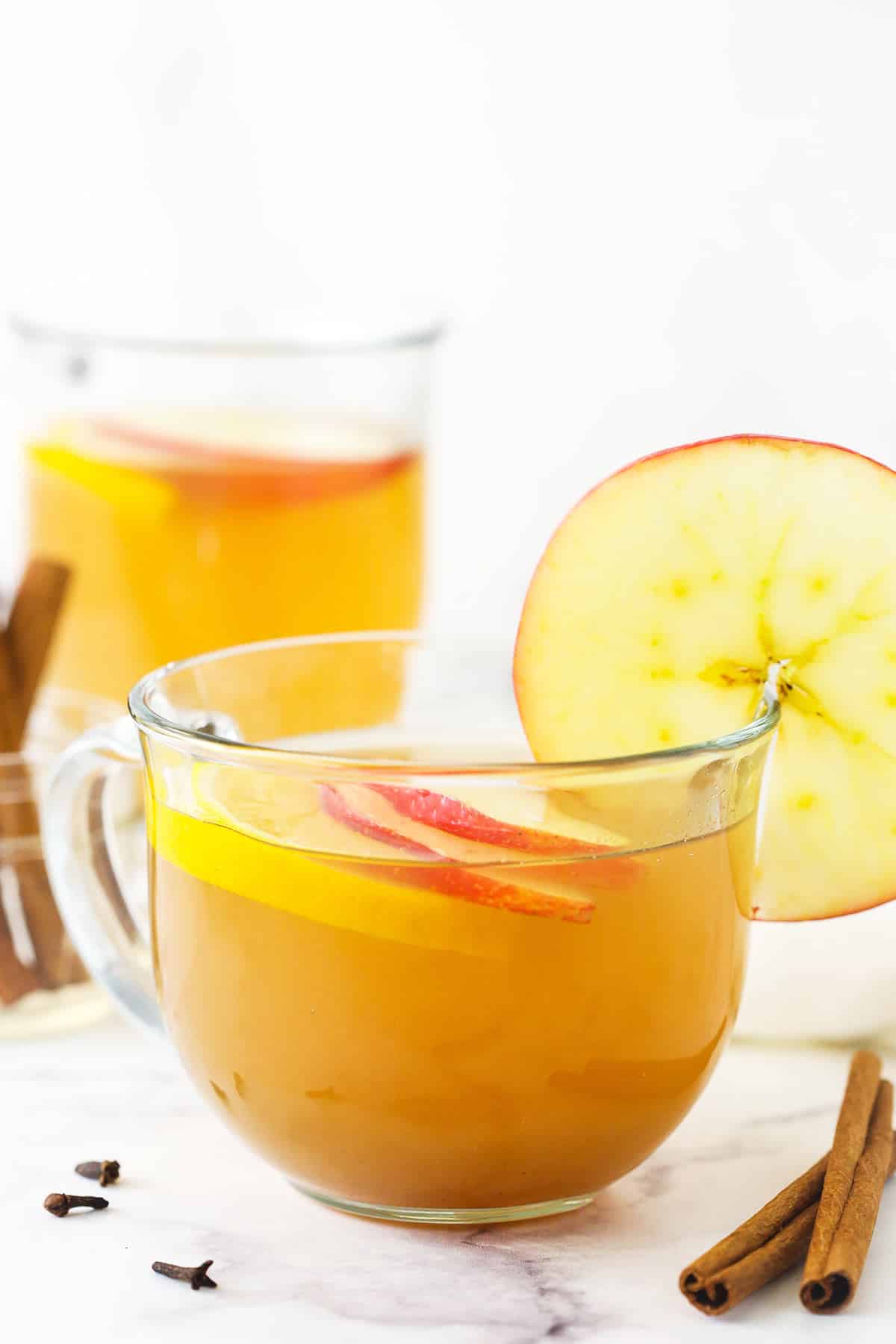 A small mug full of hot apple cider with two cinnamon sticks on the counter beside it