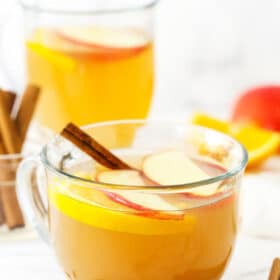 A mug of homemade apple cider with a jar of cinnamon sticks and another mug of cider behind it