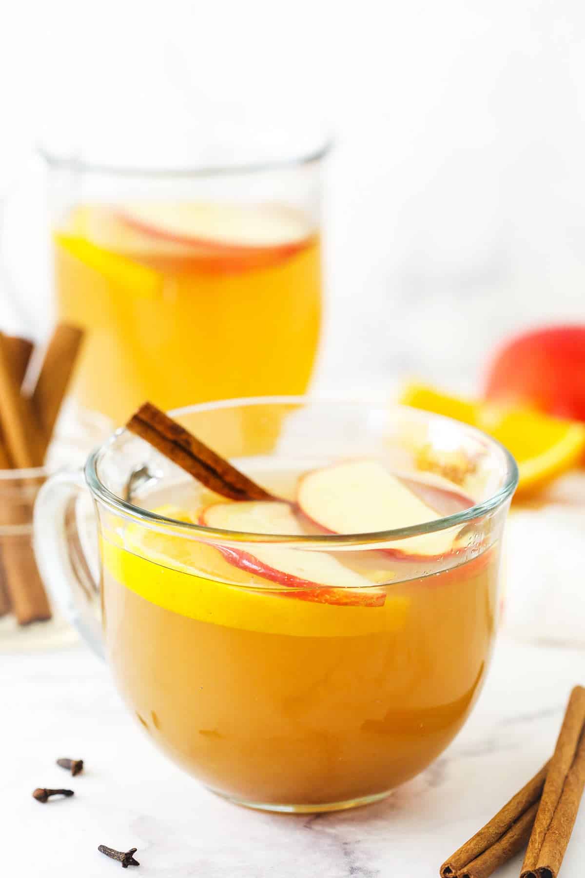 A mug of hot apple cider with a jar of cinnamon sticks and another mug of cider behind it