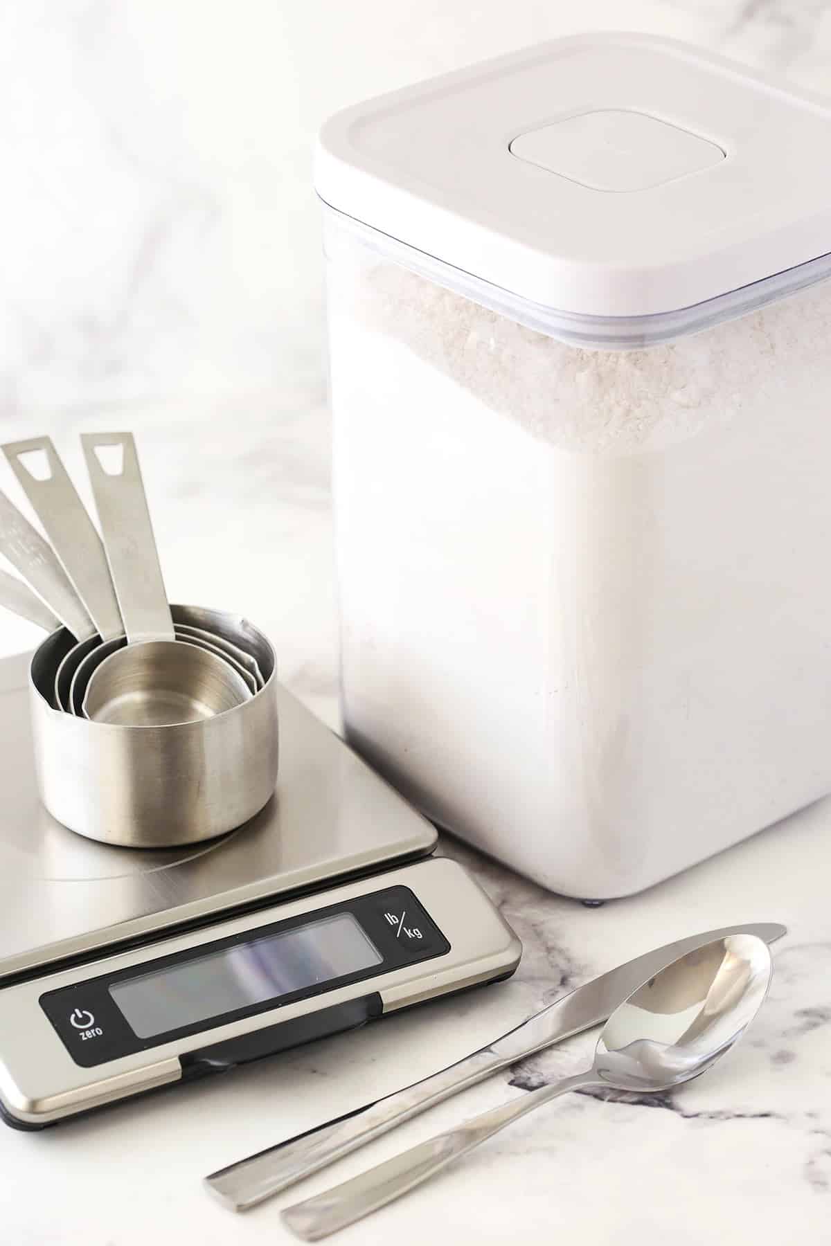 A container full of flour beside a food scale, a set of measuring cups, a fork and a butter knife
