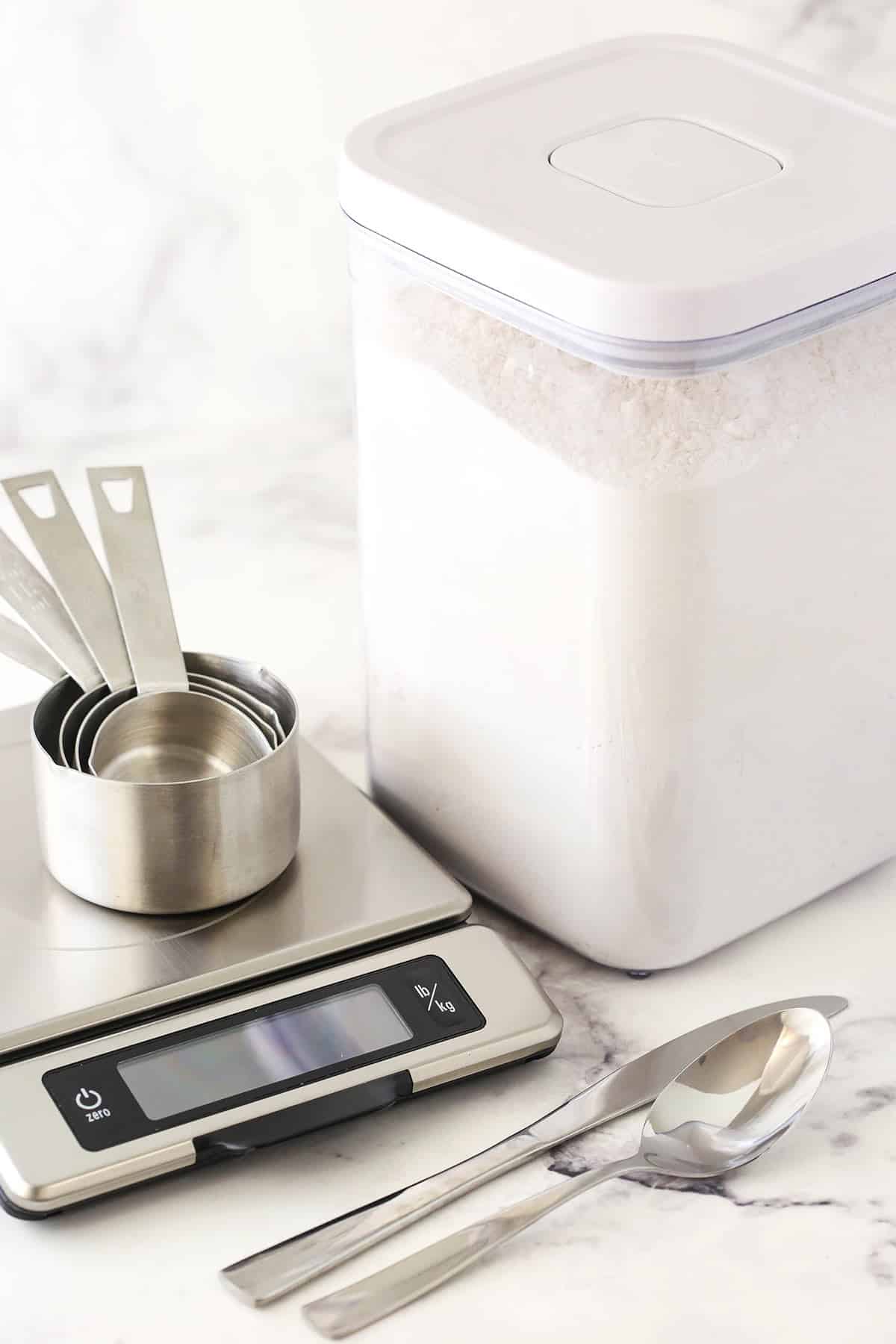Mismeasured flour can lead to dense cakes, dry cookies and cakey brownies. Learn How to Measure Flour Accurately with this helpful tutorial! A container full of flour beside a food scale, a set of measuring cups, a fork and a butter knife