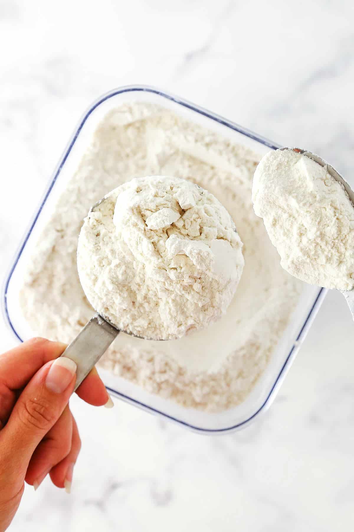 A spoon full of flour hovering over a container of flour beside a full measuring cup