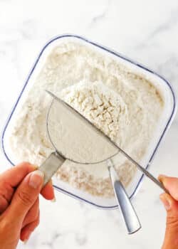 The blunt end of a butter knife being used to level off the flour overflowing from a measuring cup