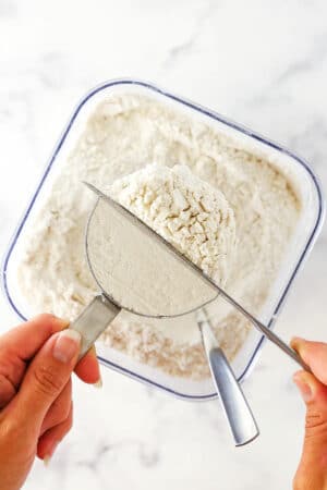 The blunt end of a butter knife being used to level off the flour overflowing from a measuring cup