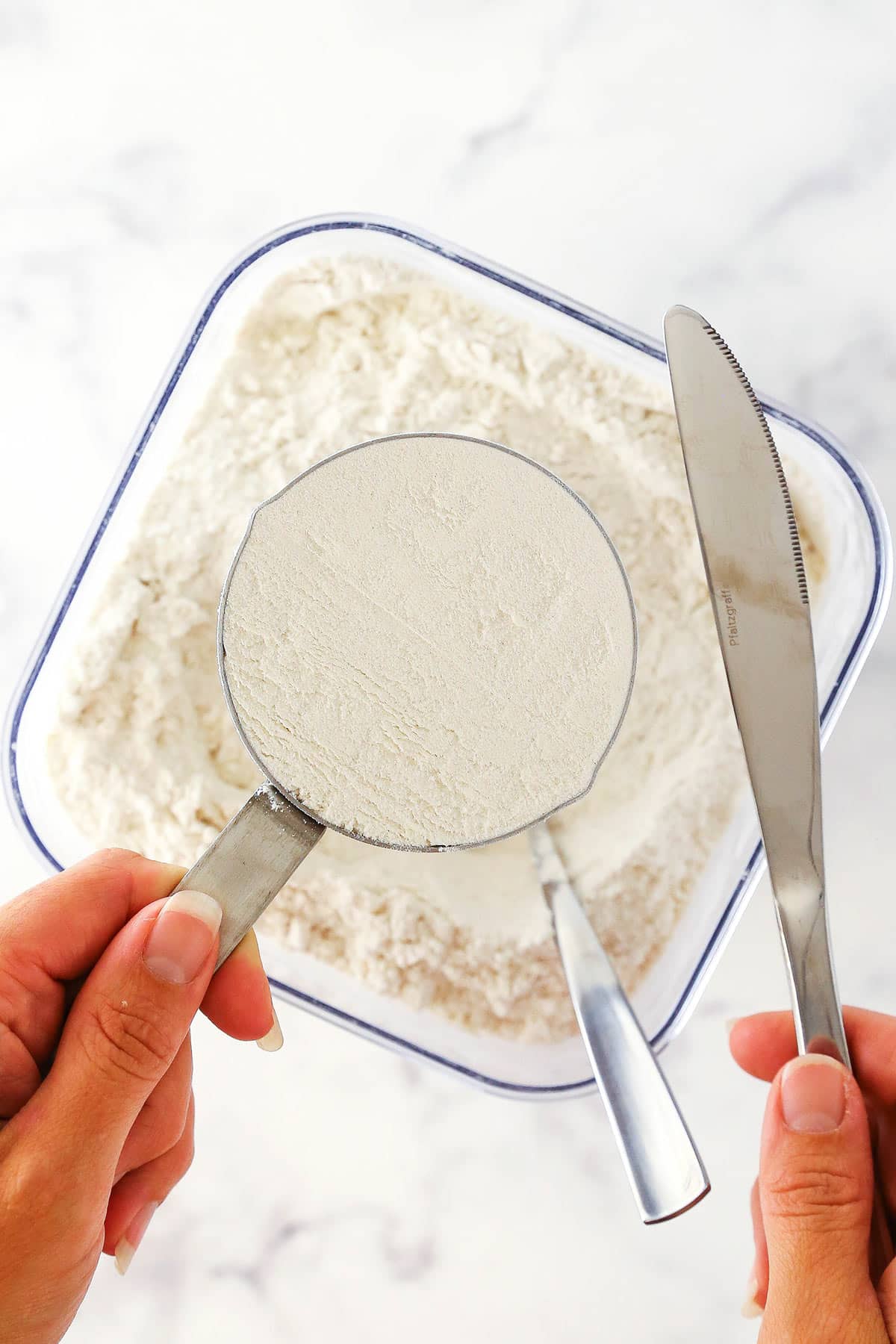 A butter knife and a cup of all-purpose flour being held above a container full of flour