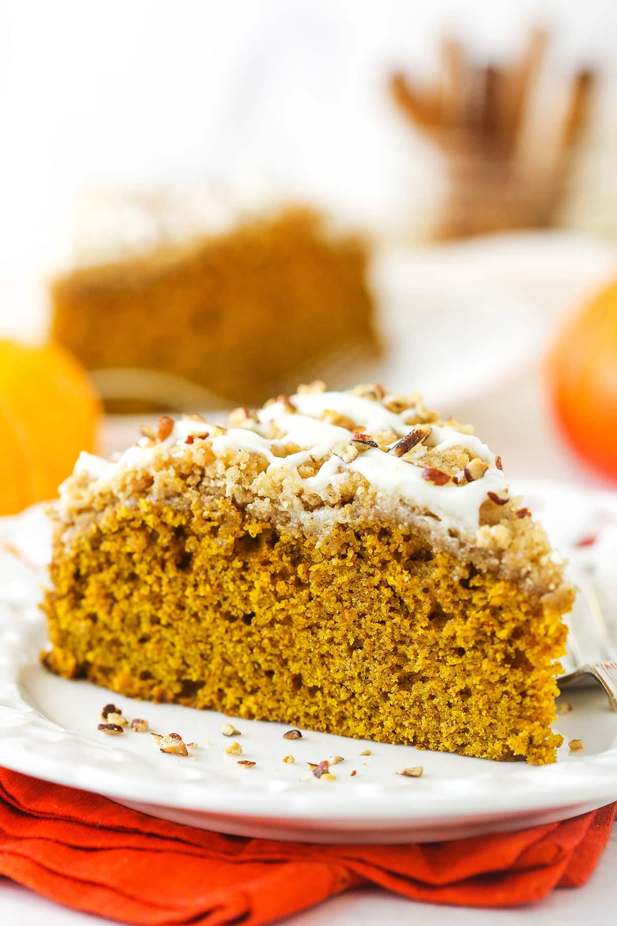 A slice of pumpkin coffee cake on a plate with a second slice and a jar of cinnamon sticks in the background.