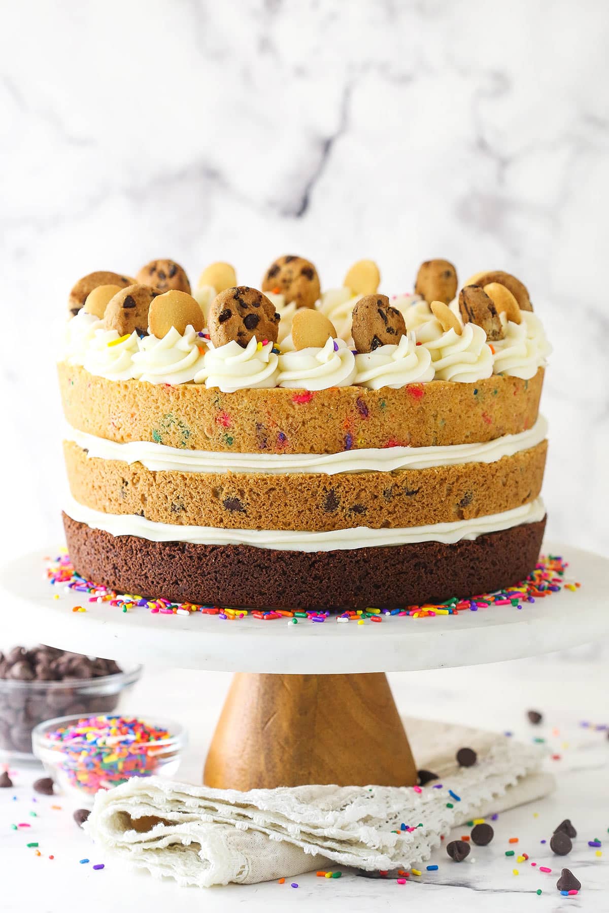 A double chocolate, chocolate chip and Funfetti cookie cake on a tall metal cake stand on top of a fancy cloth napkin with a lace trim