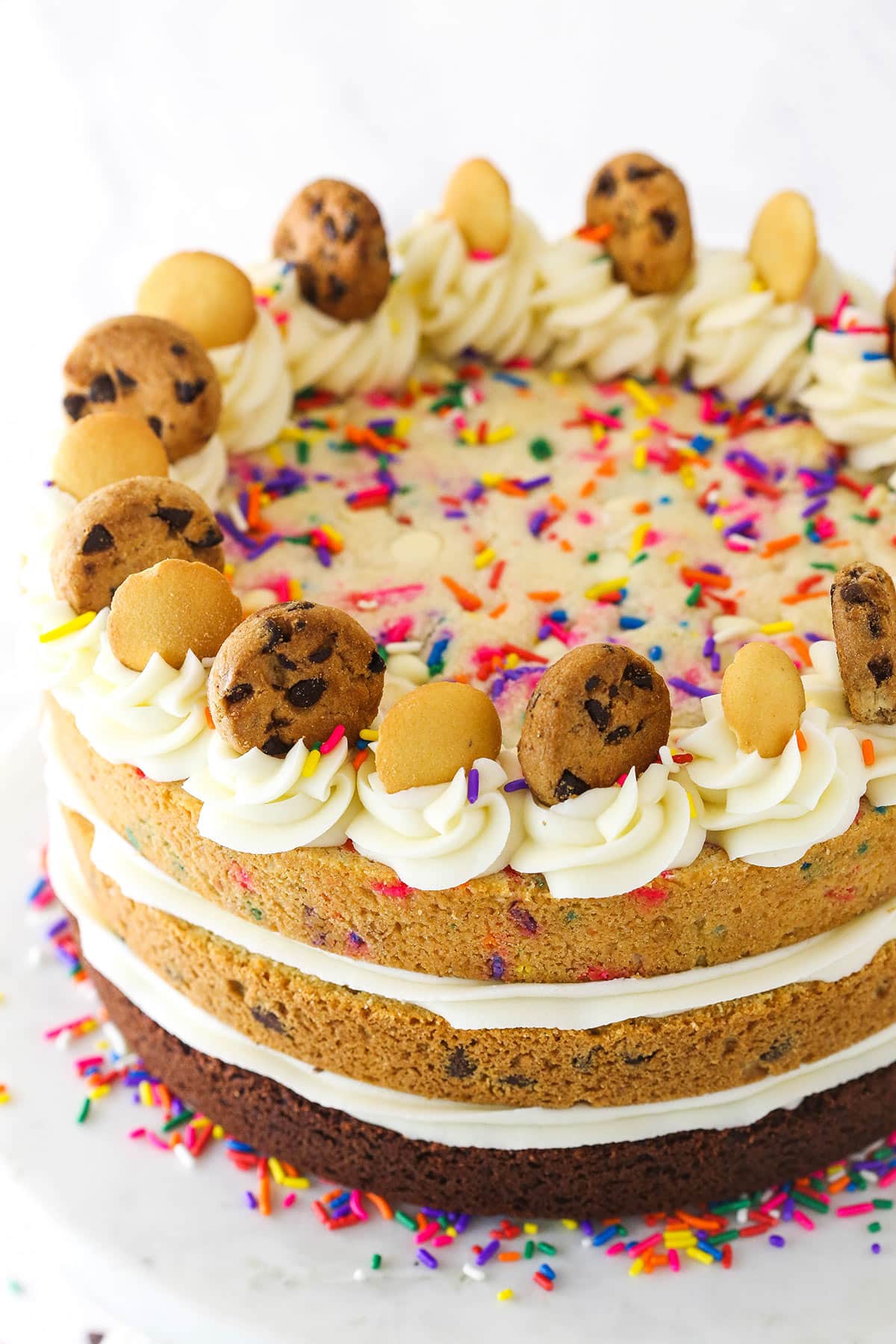 A triple-layer cookie cake on a cake stand with sprinkles and mini Nilla wafers and mini chocolate chip cookies on top