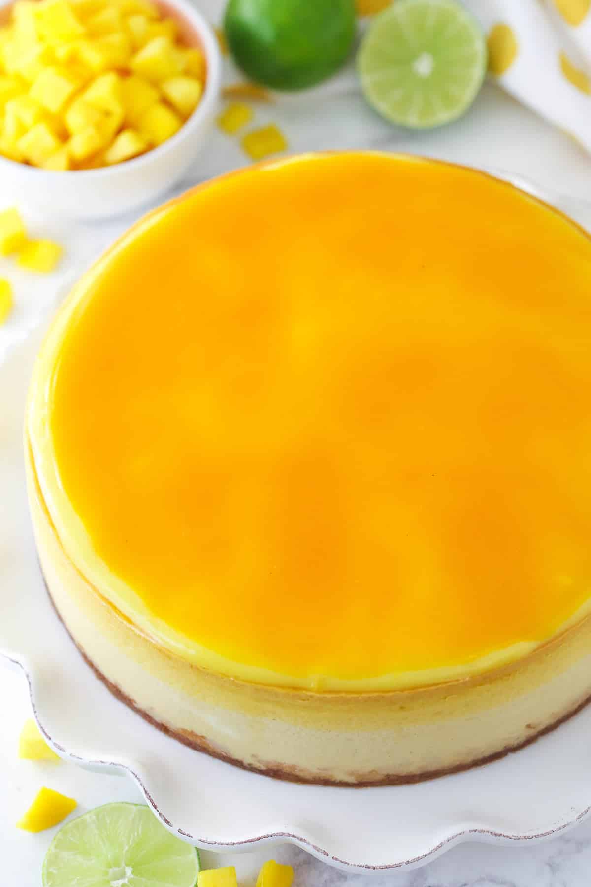 full mango key lime cheesecake on ruffled white platter with chopped mango limes in the background