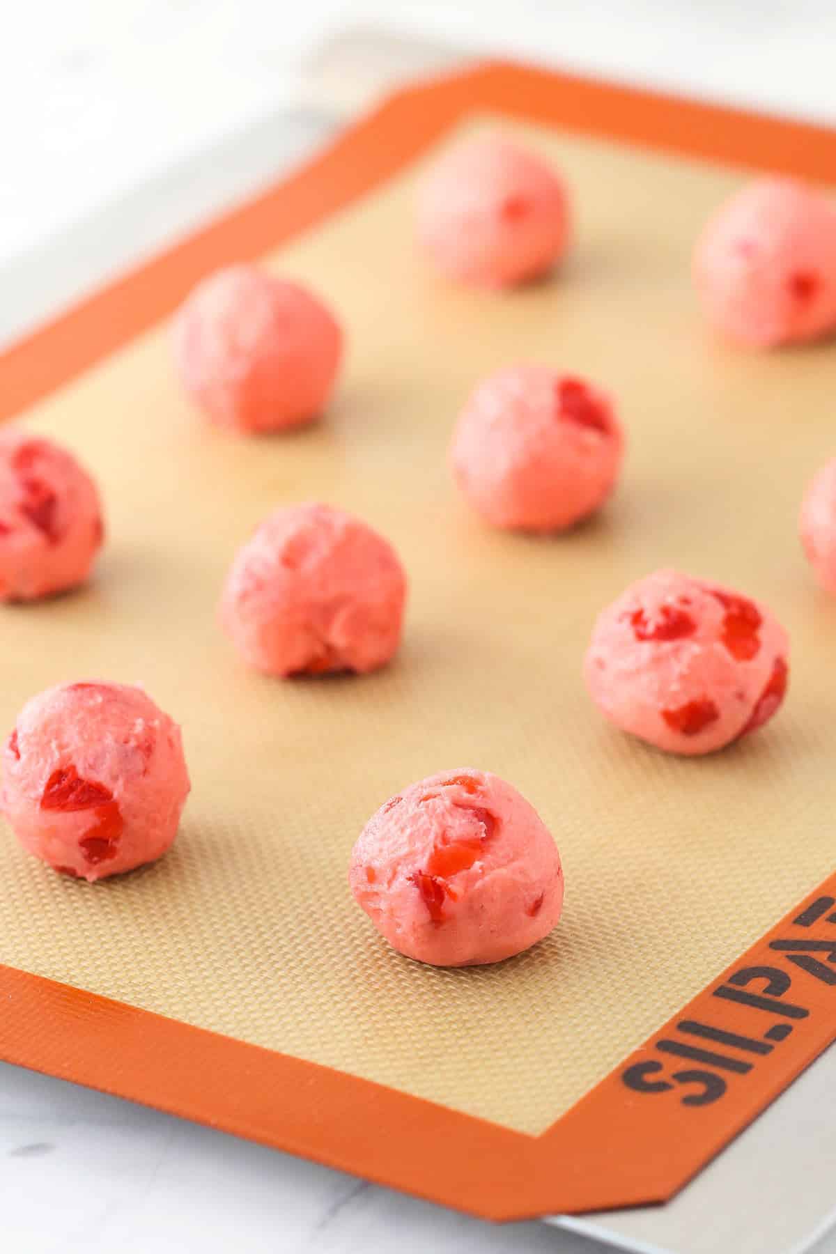 Wonderfully cute, these cherry chocolate kiss cookies are the perfect holiday treat. The secret to their flavor? Cherry juice. Balls of dough on a silicone baking sheet.