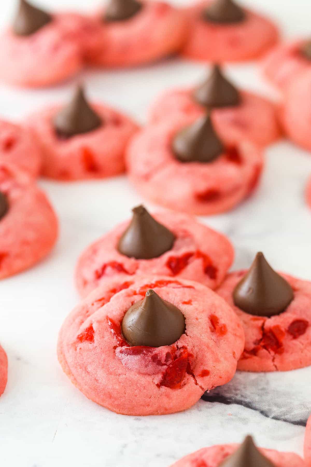 Wonderfully cute, these cherry chocolate kiss cookies are the perfect holiday treat. The secret to their flavor? Cherry juice. Lots of cherry kiss cookies on a flat surface.