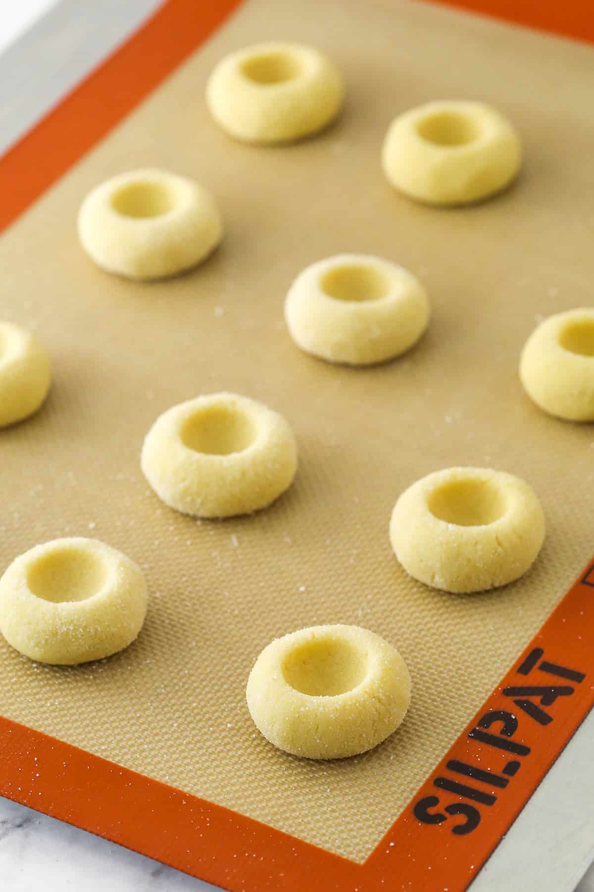 Cookie dough balls on a baking sheet and silicone baking mat with the centers pressed down.