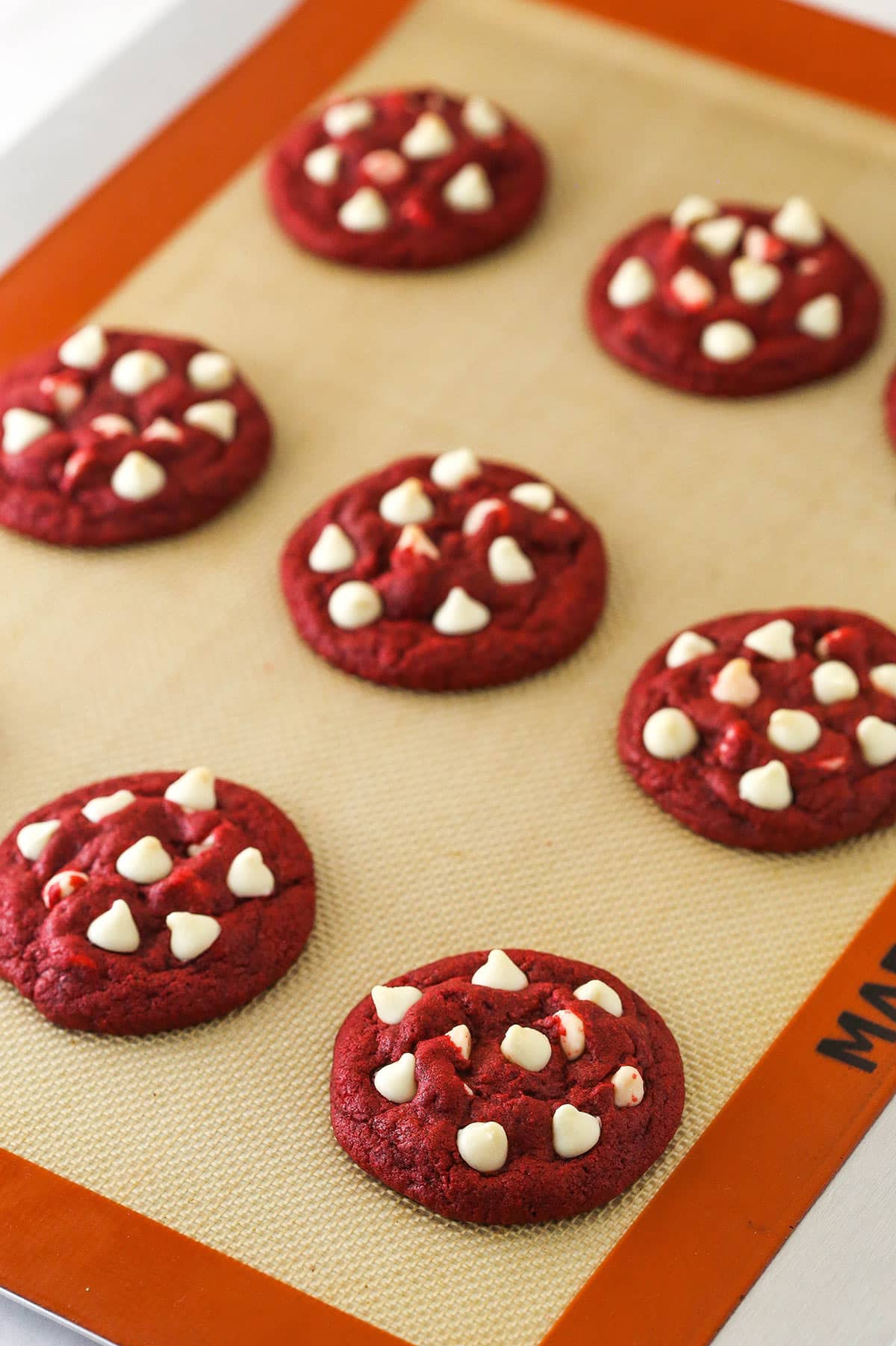 Baked red velvet cookies with white chocolate chips on a silicone baking mat.
