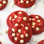 Angled shot of three red velvet cookies with white chocolate chips, with more cookies in the background.