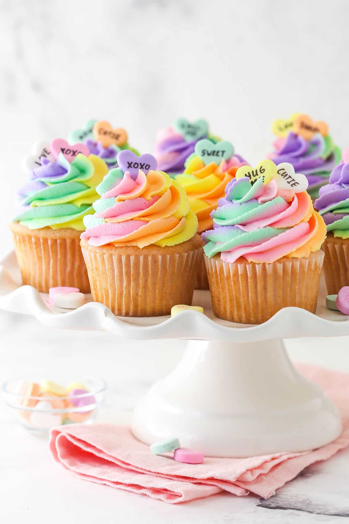 Conversation heart cupcakes arranged on a cupcake stand with conversation heart candies scattered around.