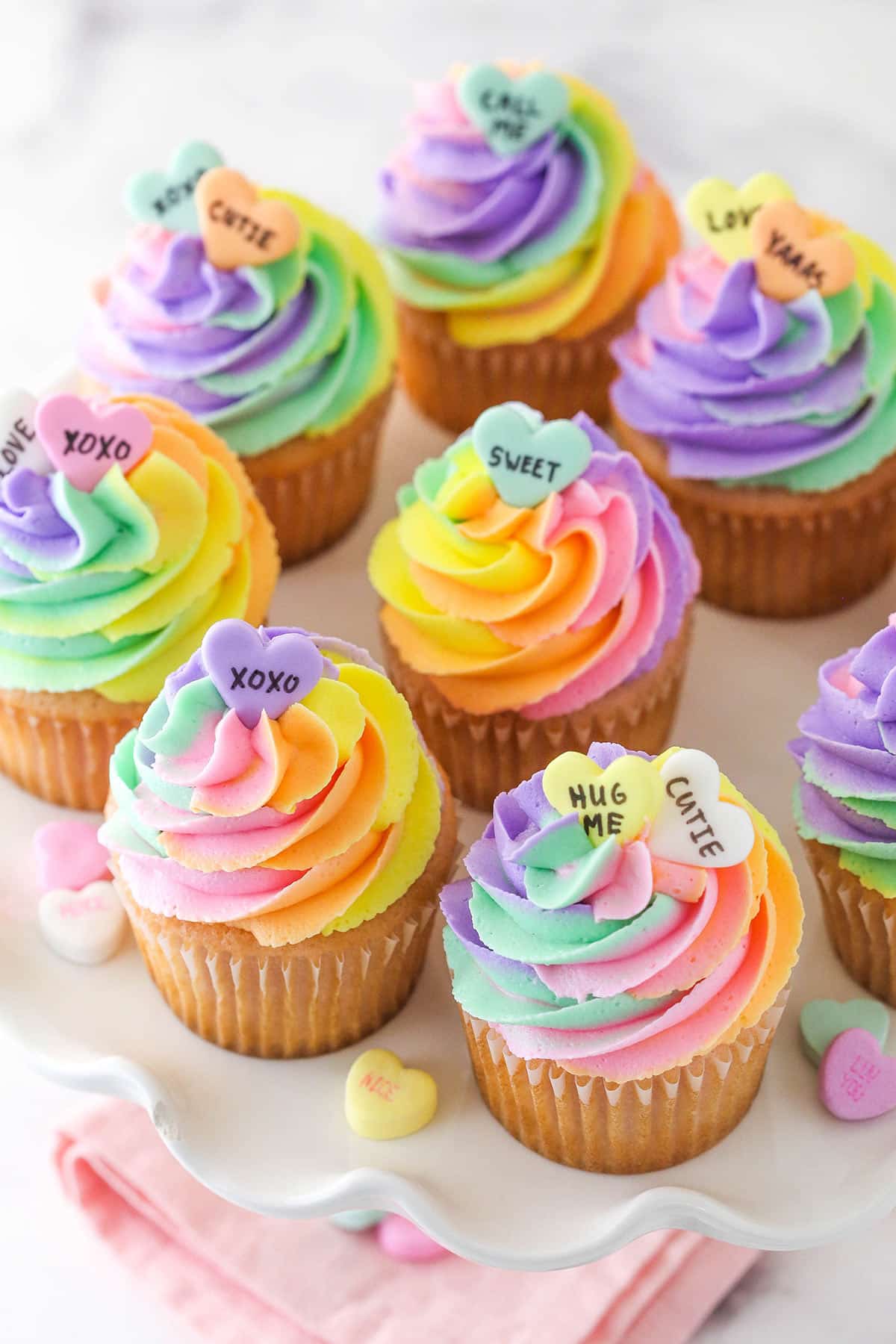 Conversation heart cupcakes arranged on a cupcake stand with conversation heart candies scattered around.