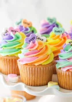 Conversation heart cupcakes arranged on a cupcake stand with conversation heart candies scattered around.