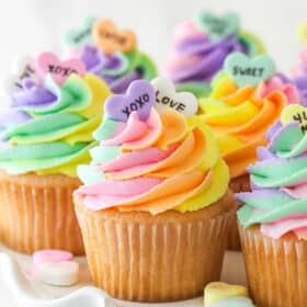 Conversation heart cupcakes arranged on a cupcake stand with conversation heart candies scattered around.