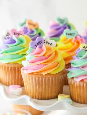 Conversation heart cupcakes arranged on a cupcake stand with conversation heart candies scattered around.