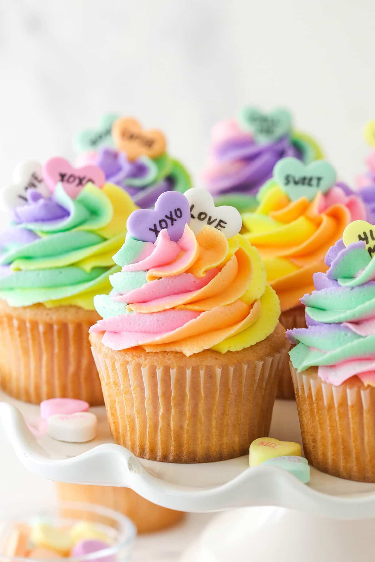 Conversation heart cupcakes arranged on a cupcake stand with conversation heart candies scattered around.