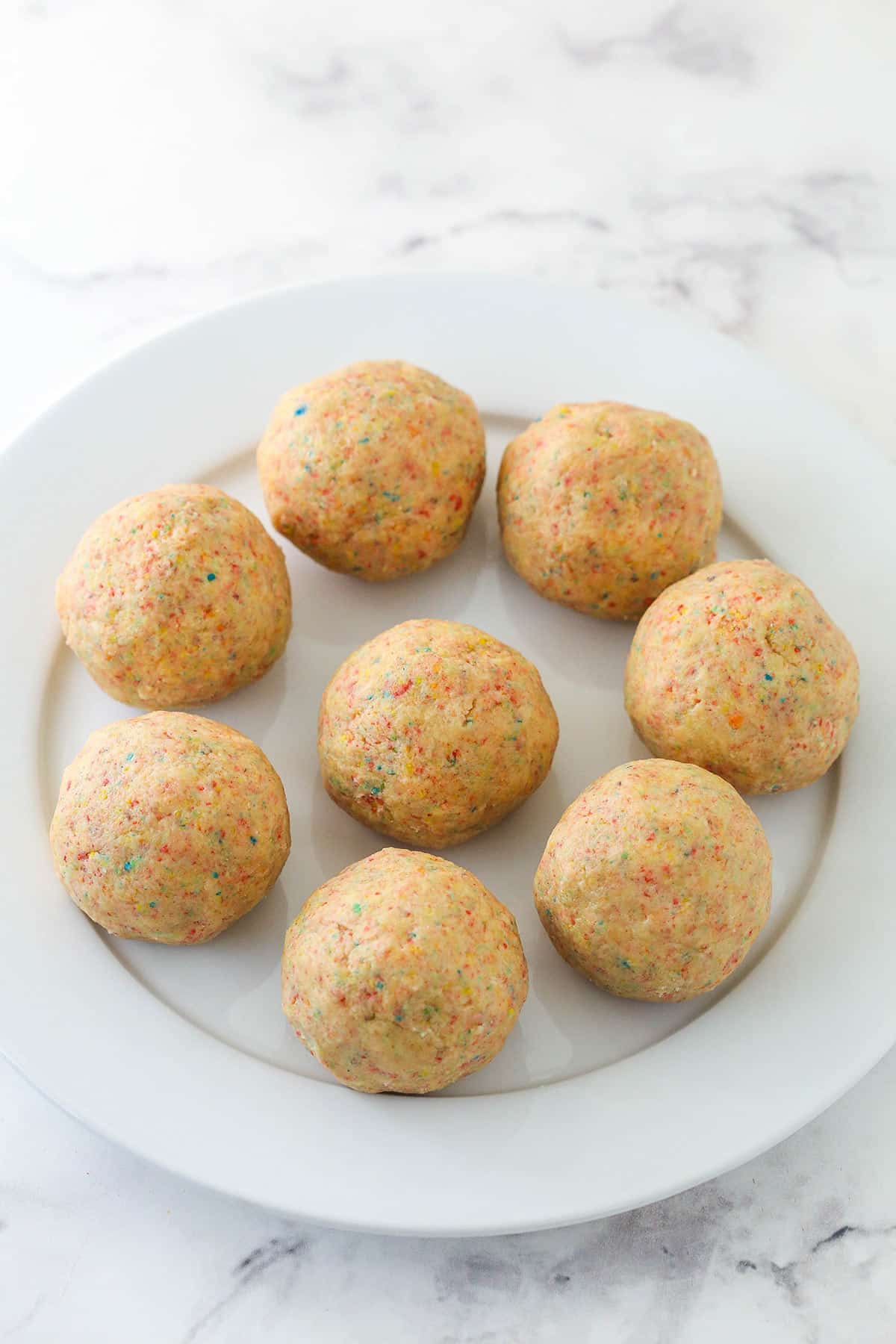 Chewy breakfast cereal infused cookies are coated in a light glaze and dipped in bright Fruity Pebbles to make rainbow Fruity Pebbles Cookies. Dough for Fruity Pebbles cookies rolled into balls and arranged on a plate.