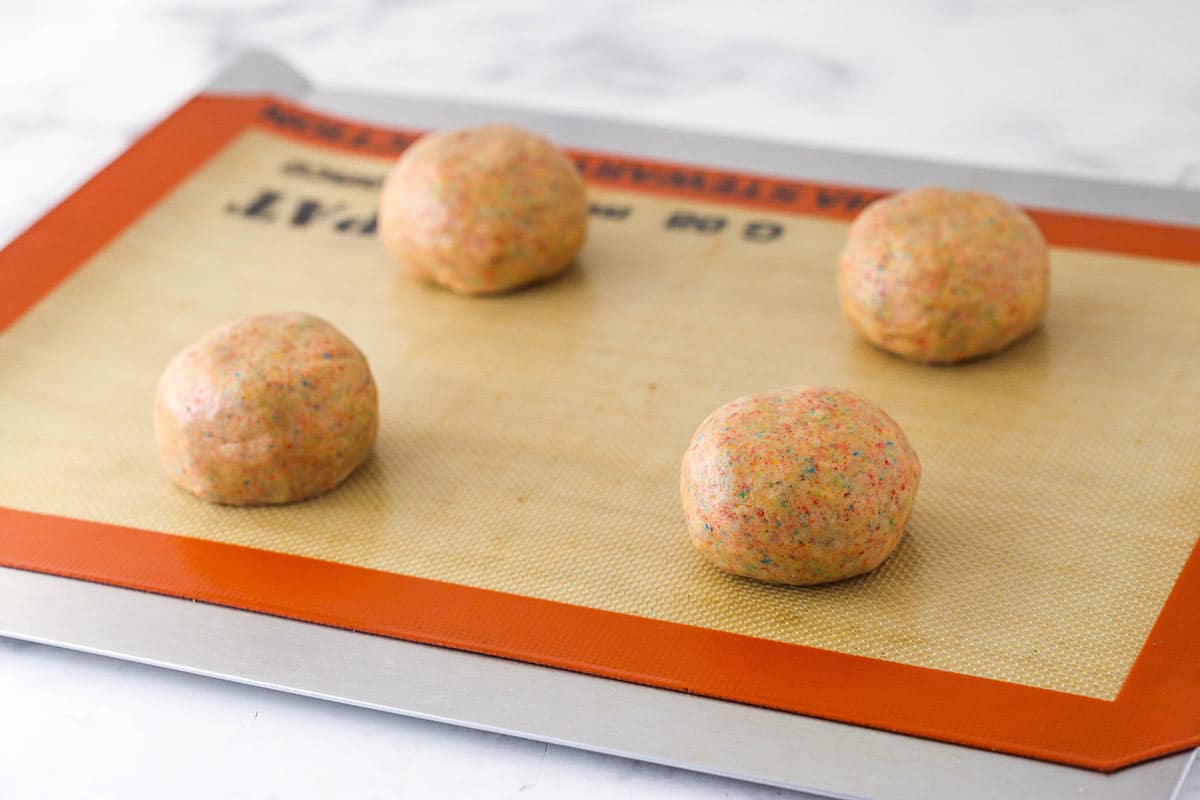 Dough for Fruity Pebble cookies rolled into balls and arranged on a silicone mat over a cookie sheet ready to be baked.