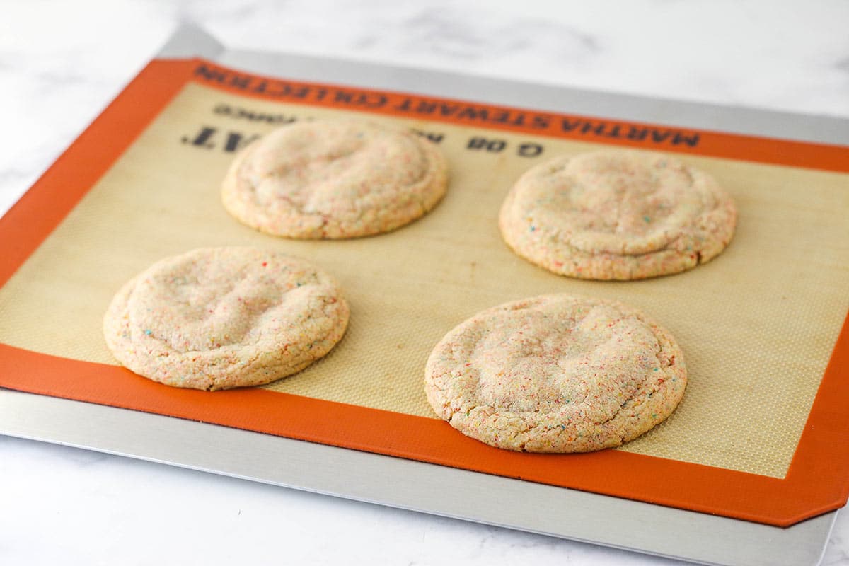 Chewy breakfast cereal infused cookies are coated in a light glaze and dipped in bright Fruity Pebbles to make rainbow Fruity Pebbles Cookies. Fruity Pebbles cookies cooling on a silicone mat.